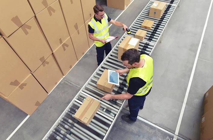 Two warehouse workers in high-visibility vests sorting and scanning packages on a conveyor belt, illustrating efficient logistics processes for Amazon FBA.